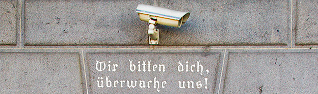 Überwachungskamera an der Hausmauer mit der Schrift „Wir bitten dich, überwache uns” darunter